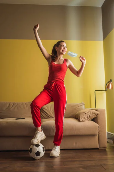 Deportista alegre con la mano por encima de la cabeza tocando máscara médica y de pie cerca del fútbol en la sala de estar, fin del concepto de cuarentena — Stock Photo