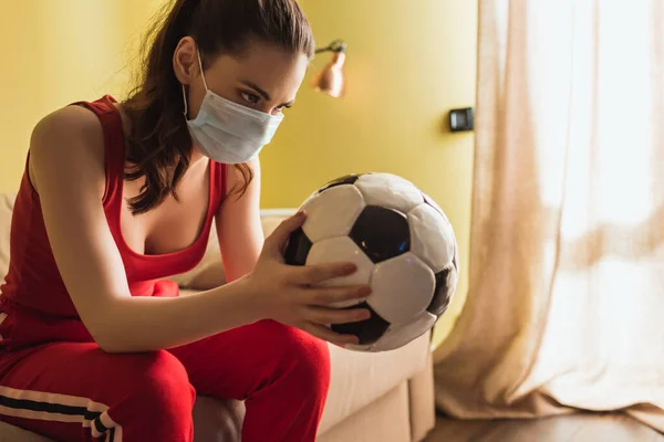 Mulher esportiva em máscara médica olhando para o futebol na sala de estar — Fotografia de Stock