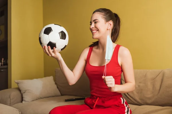 Glückliche Sportlerin berührt medizinische Maske und hält Fußball, Ende der Quarantäne — Stockfoto