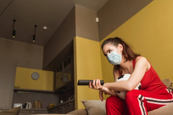 Sportswoman in medical mask holding remote controller near football while watching championship in living room — Stock Photo