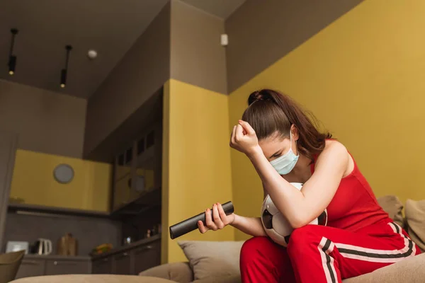 Verärgerte Sportlerin mit medizinischer Maske hält Fernbedienung in der Nähe des Fußballs, während sie die WM im Wohnzimmer verfolgt — Stockfoto