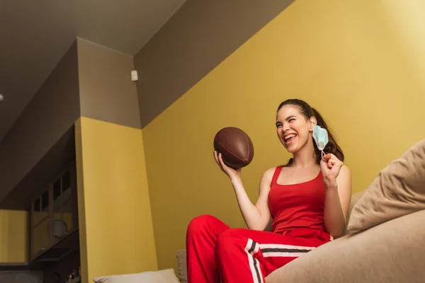 Cheerful sportswoman touching medical mask and holding american football, end of quarantine concept — Stock Photo