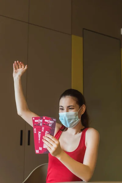 Счастливая женщина в медицинской маске, держащая ночные билеты на вечеринку дома — стоковое фото