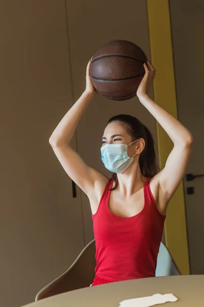 Sportive woman in medical mask holding basketball above head — Stock Photo