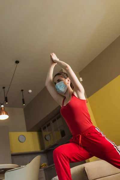 Visión de ángulo bajo de la mujer deportiva en máscara médica y la mano por encima de la cabeza haciendo ejercicio en casa - foto de stock