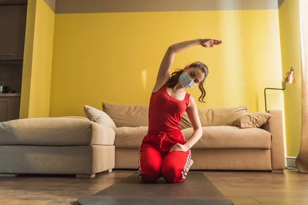 Giovane donna in maschera medica e mano sull'anca che esercita sul tappeto fitness — Foto stock