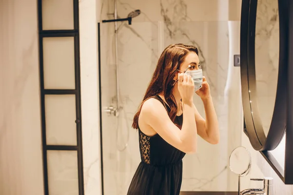 Selective focus of woman in black dress and medical mask applying eye shadow with cosmetic brush in bathroom — Stock Photo