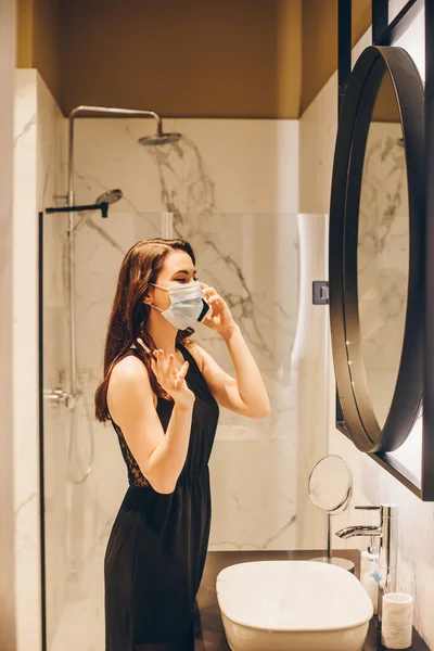 Donna in abito nero e maschera medica tenendo pennello cosmetico e parlando su smartphone in bagno — Foto stock