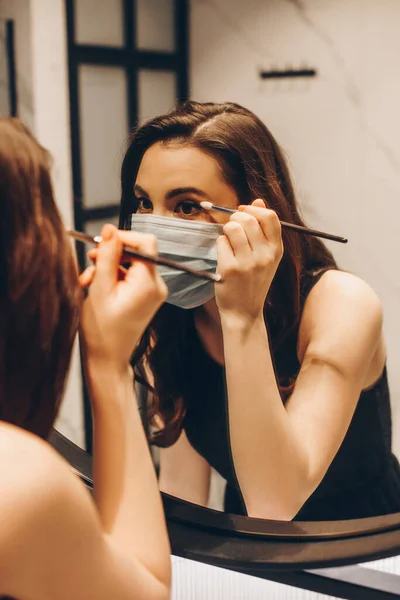 Selective focus of woman in medical mask and black dress applying eye shadow in bathroom — Stock Photo