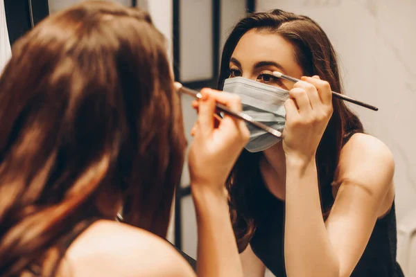 Selective focus of young woman in medical mask and black dress applying eye shadow in bathroom — Stock Photo