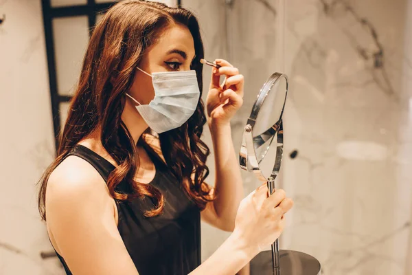 Donna in maschera medica e vestito nero applicando ombretto e tenendo specchio in bagno — Foto stock