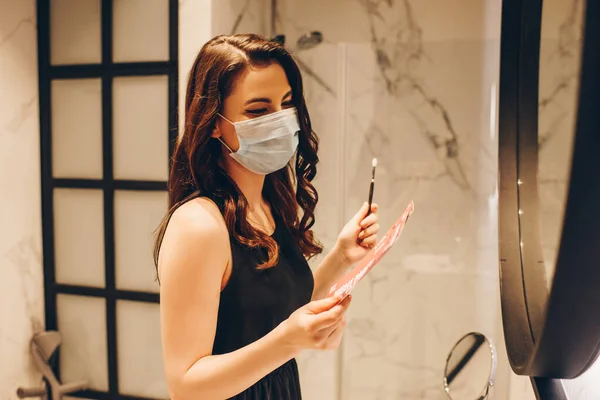 Donna in maschera medica e vestito nero con biglietti per la festa notturna e pennello cosmetico, fine del concetto di quarantena — Foto stock