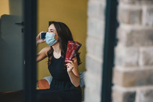 Selective focus of young woman in medical mask and black dress holding night party tickets and talking on smartphone, end of quarantine concept — Stock Photo