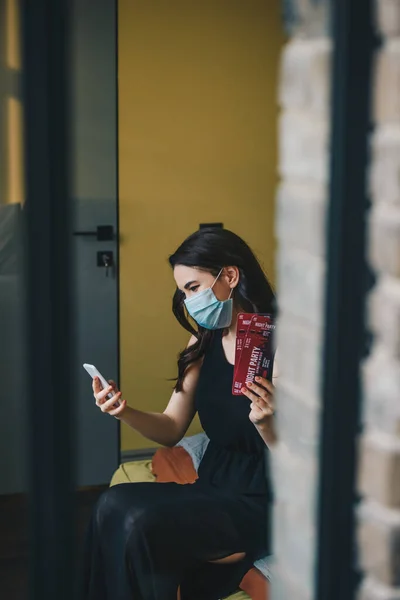 Selective focus of young woman in medical mask and black dress holding night party tickets and using smartphone, end of quarantine concept — Stock Photo