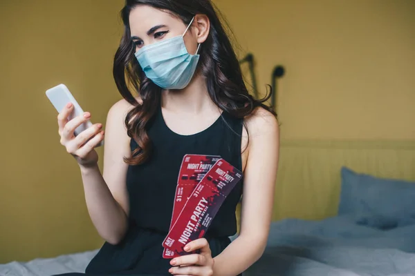 Young woman in medical mask and black dress holding night party tickets and using smartphone, end of quarantine concept — Stock Photo