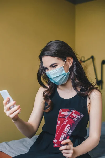 Enfoque selectivo de la niña en máscara médica celebración de entradas de fiesta de noche y el uso de teléfono inteligente, fin del concepto de cuarentena - foto de stock