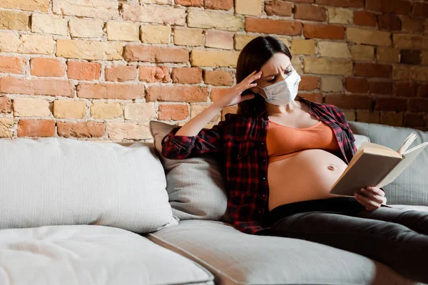 Young pregnant woman in medical mask reading book in living room — Stock Photo
