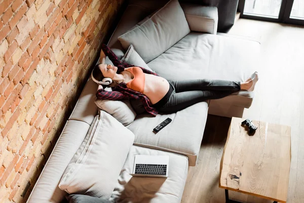 KYIV, UKRAINE - APRIL 30, 2020: overhead view of pregnant woman in headphones relaxing near laptop, joystick and remote controller on sofa — Stock Photo