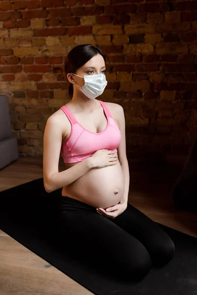 Беременная женщина в спортивной одежде и медицинской маске касаясь живота — стоковое фото