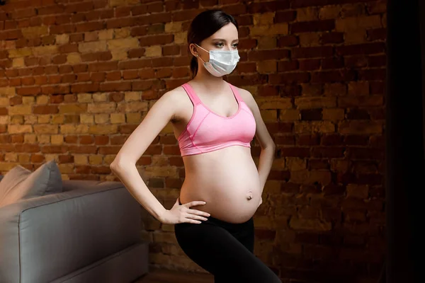 Беременная женщина в медицинской маске с руками на бедрах, тренирующимися возле кирпичной стены — стоковое фото