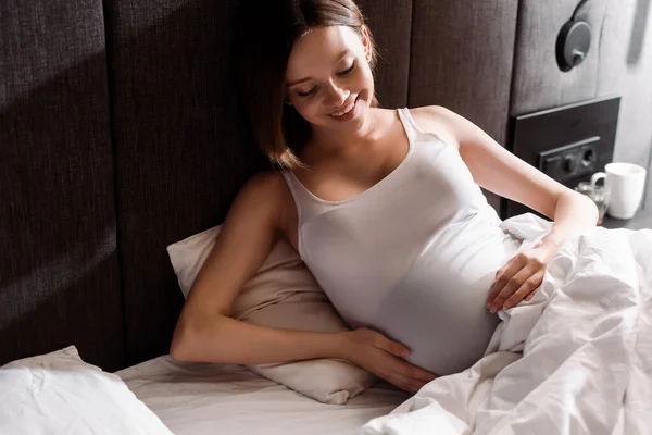 Cheerful and pregnant woman touching belly in bed — Stock Photo