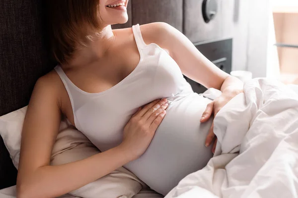 Обрезанный вид веселой и беременной женщины, касающейся живота в постели — стоковое фото