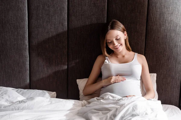 Glückliche und schwangere Frau, die Bauch im Schlafzimmer anfasst und betrachtet — Stockfoto