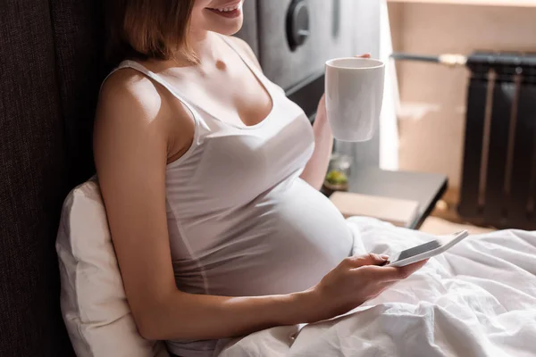 Vista ritagliata della donna incinta in possesso di tazza e utilizzando smartphone a letto — Foto stock
