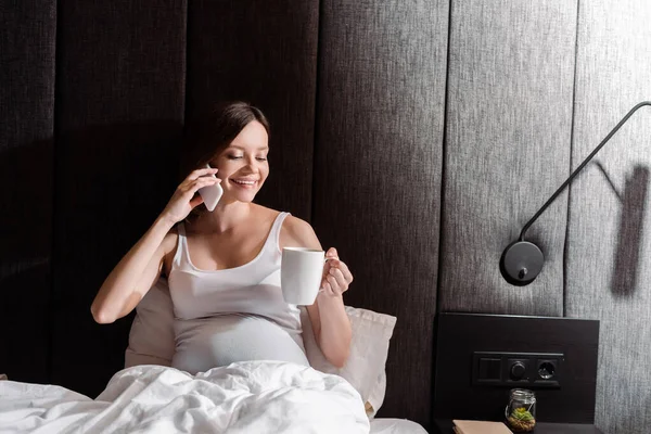 Mujer embarazada feliz sosteniendo taza y hablando en el teléfono inteligente en la cama - foto de stock