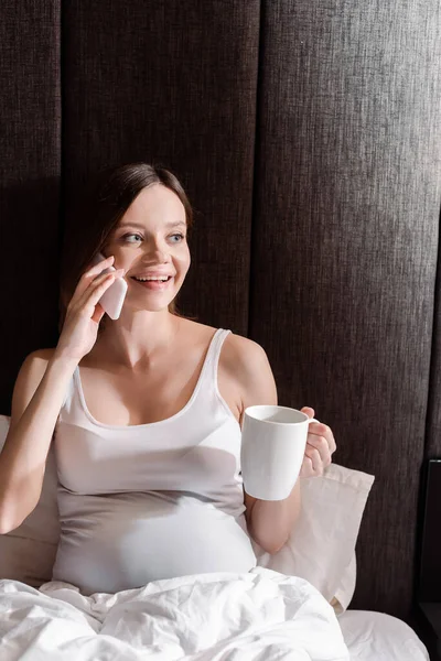 Gaie femme enceinte tenant tasse et parler sur smartphone au lit — Photo de stock