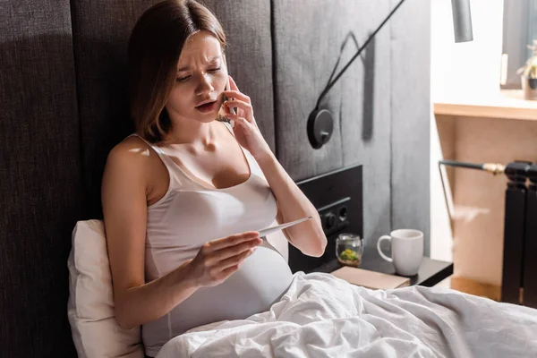 Junge schwangere Frau spricht auf Smartphone und schaut auf digitales Thermometer im Schlafzimmer — Stockfoto
