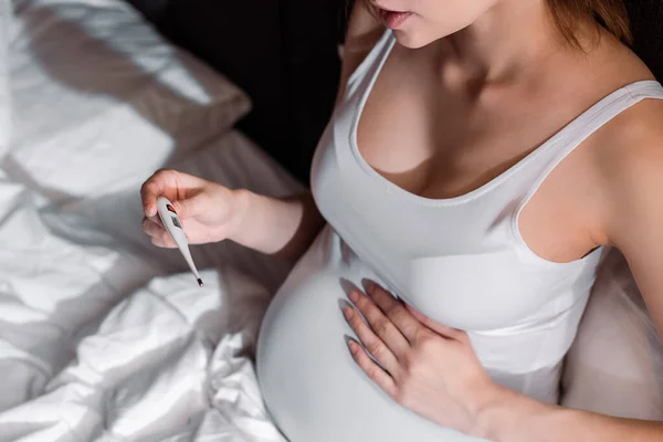 Vista ritagliata della donna incinta che tiene il termometro digitale in camera da letto — Foto stock