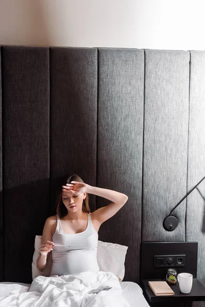 Sick pregnant woman holding digital thermometer in bedroom — Stock Photo