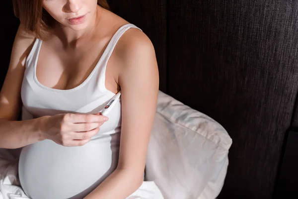 Cropped view of sick pregnant woman holding digital thermometer in bedroom — Stock Photo