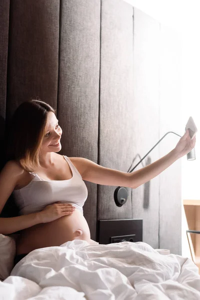 Alegre embarazada tomando selfie en dormitorio - foto de stock