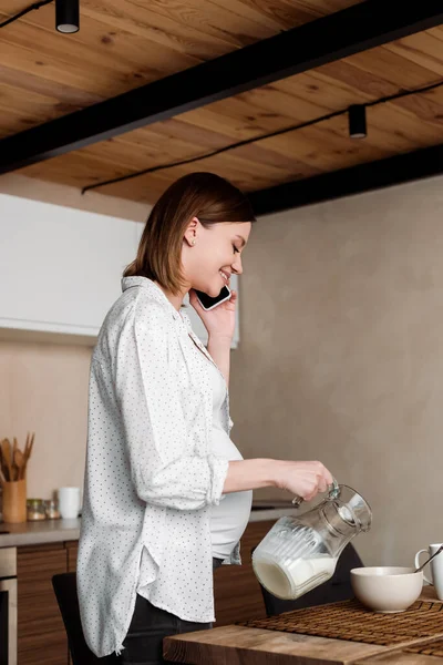 Mujer feliz y embarazada sosteniendo jarra con leche cerca del tazón y hablando en el teléfono inteligente - foto de stock
