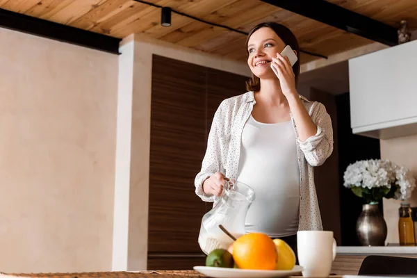 Glückliche und schwangere Frau hält Krug mit Milch in der Hand und spricht auf dem Smartphone — Stockfoto