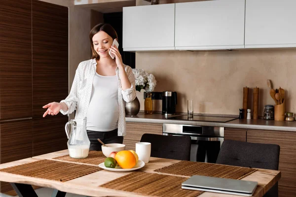 Щаслива і вагітна жінка жестикулює під час розмови на смартфоні біля їжі на кухні — стокове фото