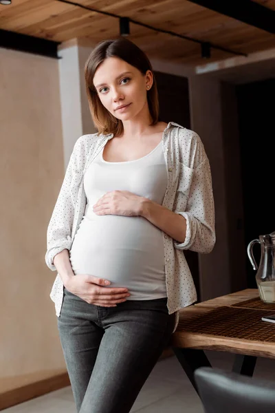 Attractive and pregnant woman touching belly while standing at home — Stock Photo