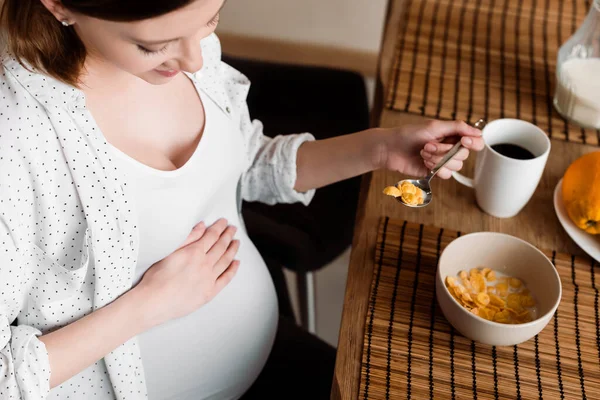 Vista aérea da mulher feliz e grávida segurando colher com saborosos flocos de milho perto da xícara com chá — Fotografia de Stock