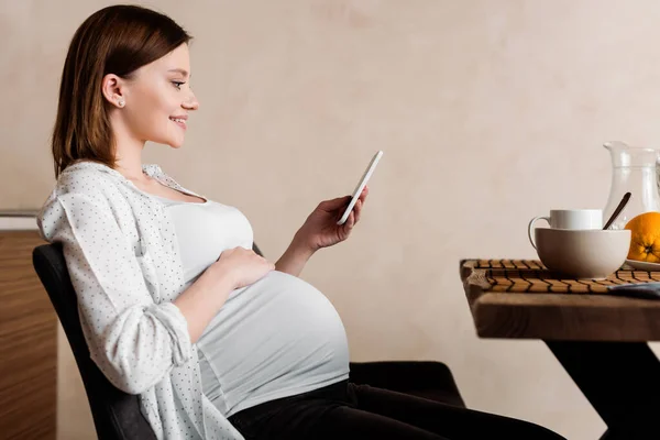 Vista lateral de la mujer embarazada feliz utilizando el teléfono inteligente cerca del desayuno sabroso - foto de stock
