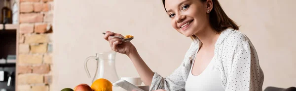 Horizontales Bild einer schwangeren Frau mit Kochlöffel mit Cornflakes und Smartphone — Stockfoto