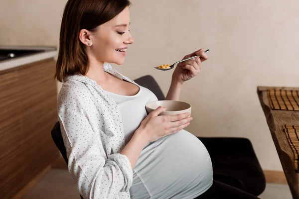 Side view of smiling pregnant woman holding spoon with corn flakes and bowl — Stock Photo