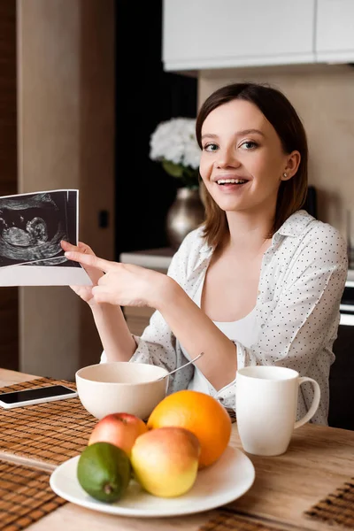 Foco seletivo de mulher alegre e grávida apontando com o dedo para fotos de ultra-som perto de frutas e smartphone com tela em branco — Fotografia de Stock