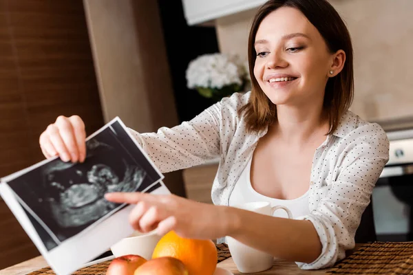Enfoque selectivo de la mujer embarazada feliz apuntando con el dedo a las fotos de ultrasonido cerca de frutas - foto de stock
