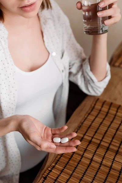 Vista recortada de la mujer embarazada sosteniendo vitaminas píldoras y vaso de agua — Stock Photo