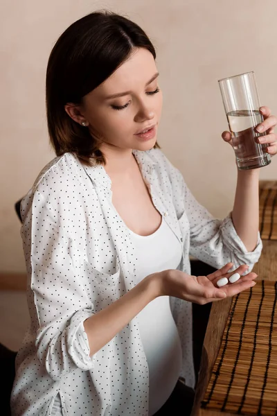 Atractiva mujer embarazada sosteniendo vitaminas píldoras y vaso de agua - foto de stock