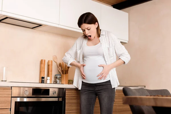 Femme enceinte toucher le ventre tout en ayant des crampes et des cris dans la cuisine — Photo de stock