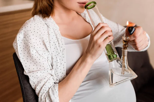 Vista recortada de una mujer embarazada sosteniendo un encendedor cerca de la pipa con cannabis legal en casa - foto de stock