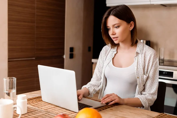 Pregnant freelancer using laptop near glass of water and bottle with vitamins — Stock Photo